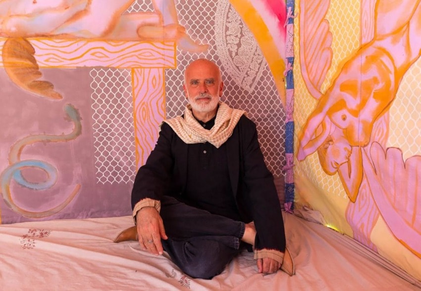 Man sitting cross-legged in a colorful room with artistic wall designs and a contemplative expression.