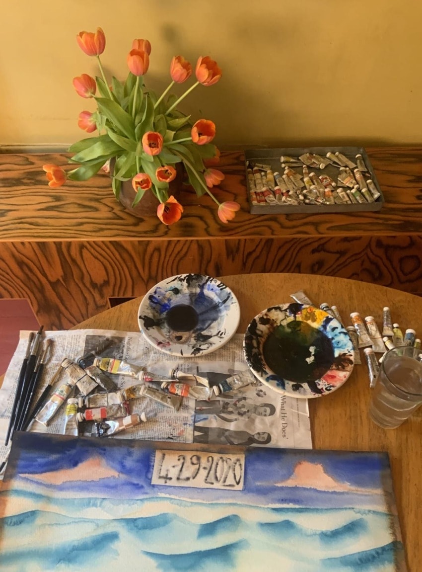 Artistic workspace with tulips, paint tubes, brushes, palettes, and a painting on a wooden table nearby with a date written.
