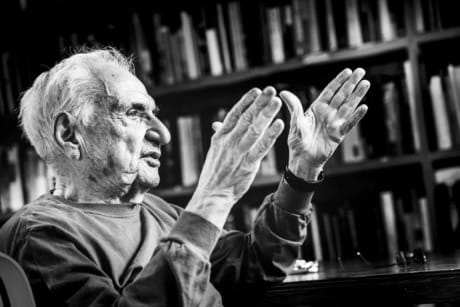 Elderly man gesturing with hands while speaking, seated in front of a bookshelf filled with books, black and white photo.
