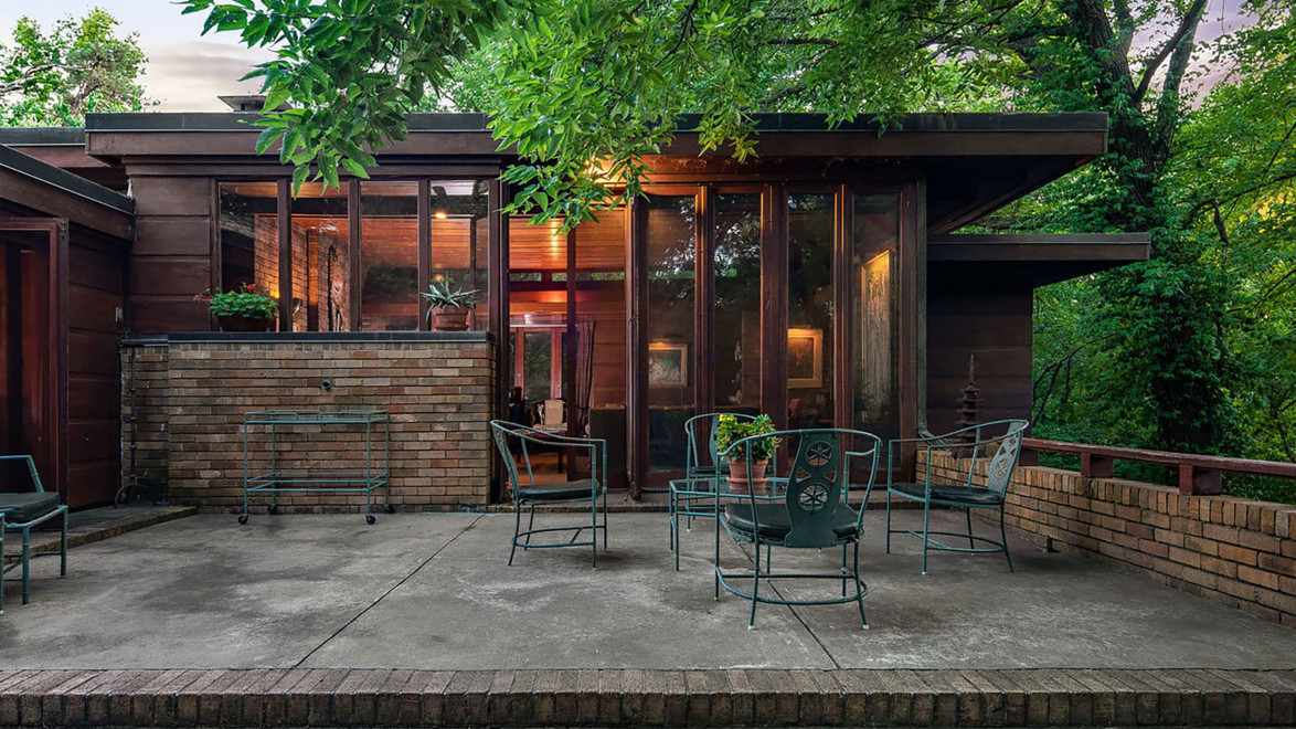 Modern house with brick and glass exterior, patio with metal chairs, surrounded by lush green trees.