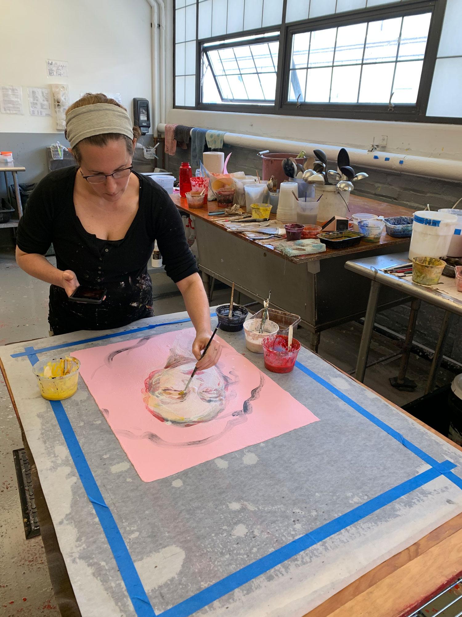 Artist painting at a table filled with art supplies in a studio.