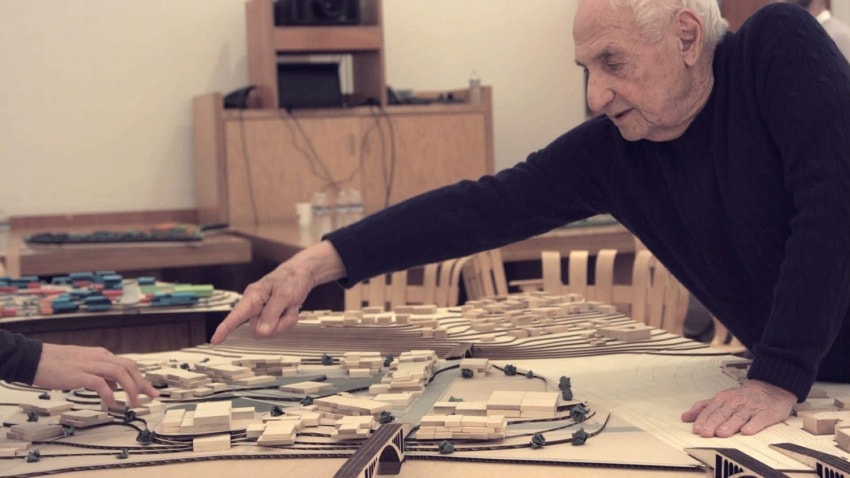 An elderly man points at an architectural model on a table surrounded by wooden chairs in a room.