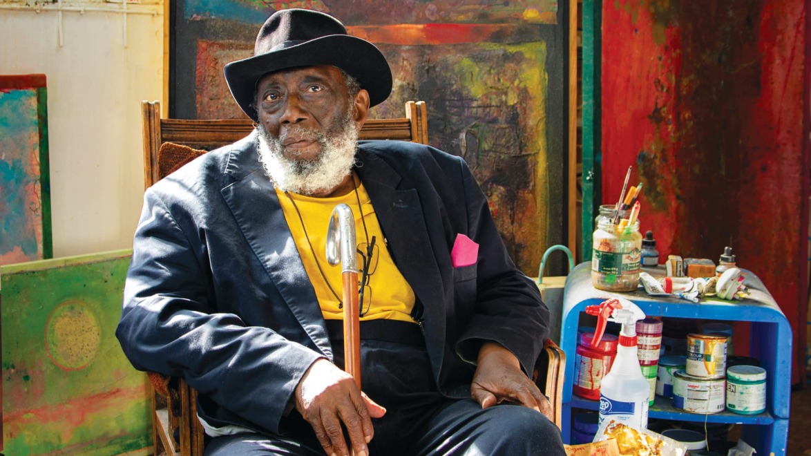 Elderly man with a beard wearing a hat and suit, seated in an art studio surrounded by colorful paintings and supplies.
