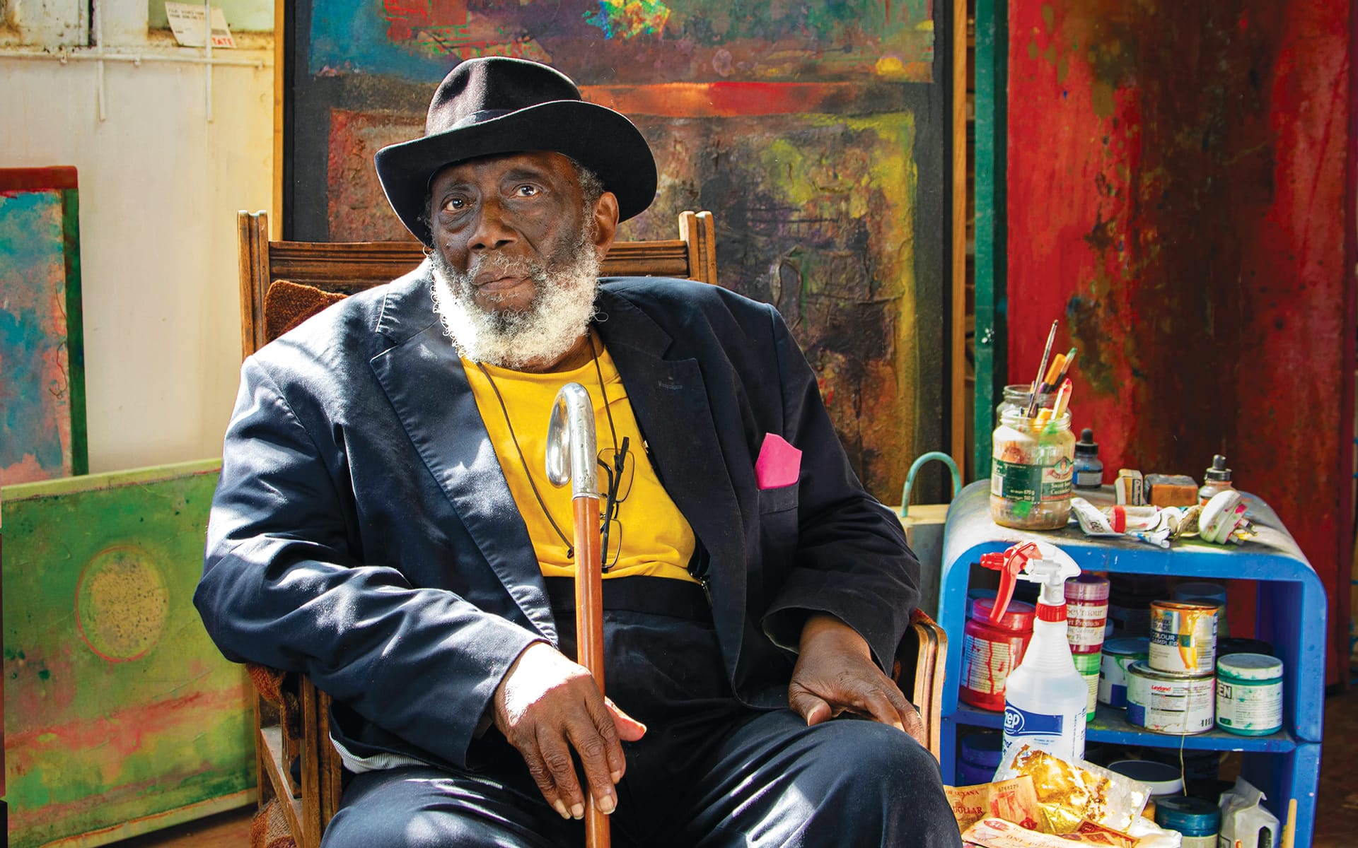 Elderly man with a beard wearing a hat and suit, seated in an art studio surrounded by colorful paintings and supplies.