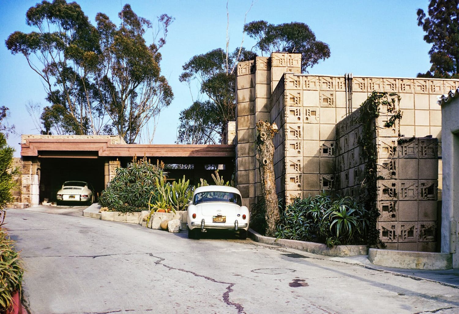 Vintage cars parked in driveway of mid-century modern house with decorative concrete blocks and surrounding greenery.