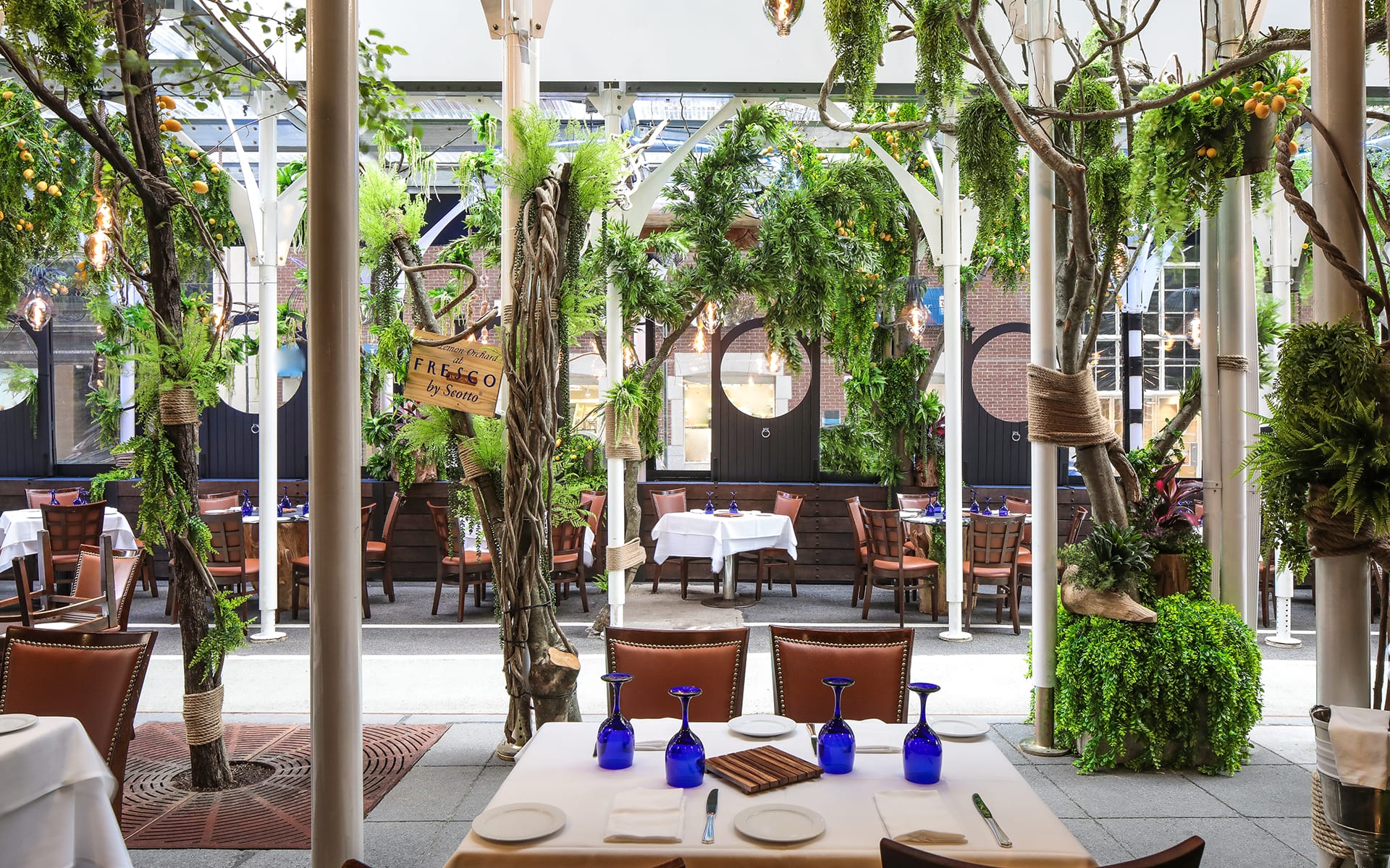 Outdoor restaurant patio with tables set, surrounded by lush greenery and decorative elements, ready for patrons.