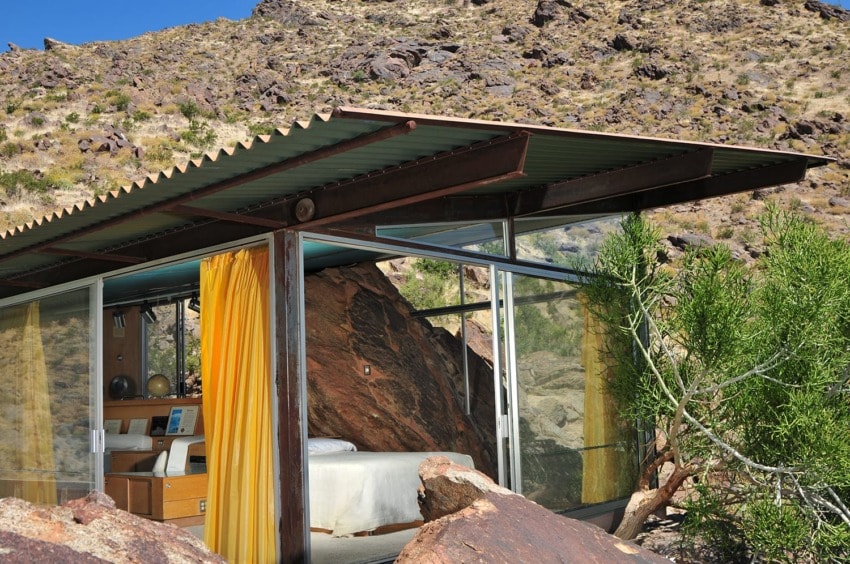 Modern glass-walled house in a desert setting with rocky landscape and yellow curtains inside.