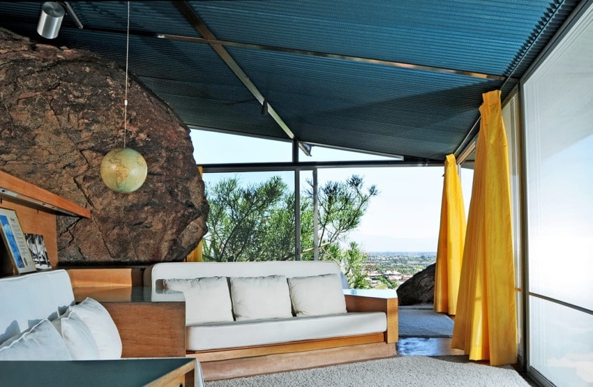 Modern living room with large windows, rock feature, yellow curtains, and a view of the city and mountains in the distance.