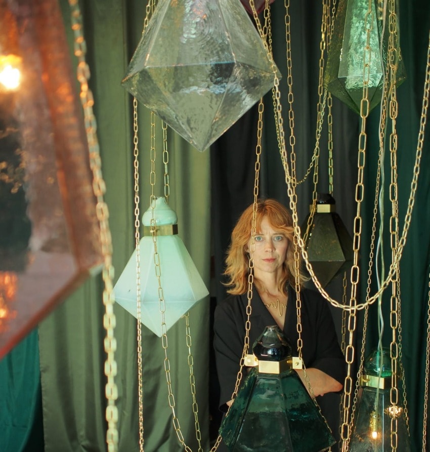 Woman stands among hanging glass lanterns and chains with a curtain backdrop.
