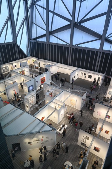 Aerial view of a modern art gallery with people exploring various exhibition booths and artworks displayed on white walls.