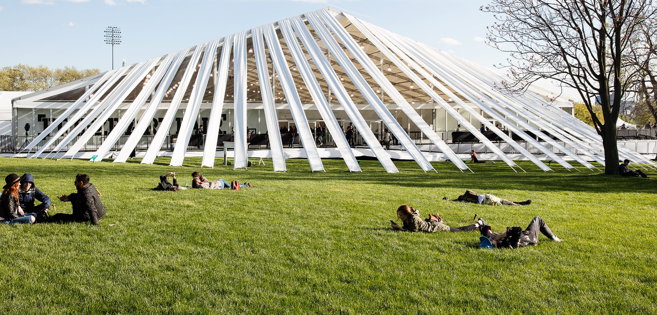 People relaxing on the grass in front of a modern, tent-like pavilion on a sunny day.