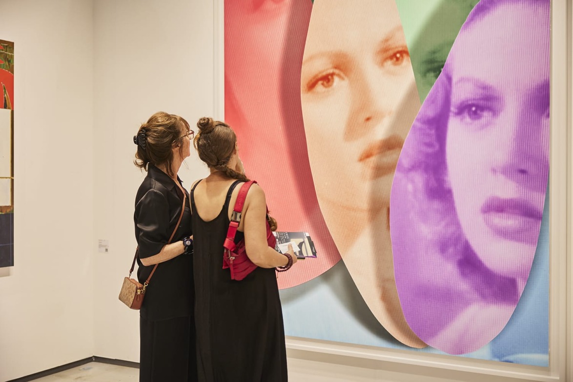 Two women observing a colorful art piece featuring faces in a modern gallery setting.
