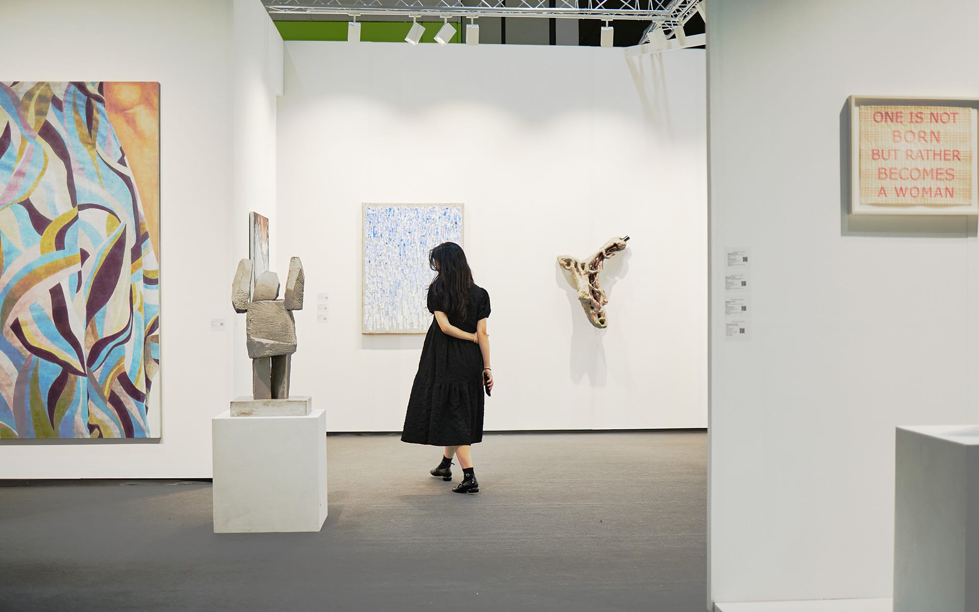 Woman in black dress viewing art in a modern gallery with various sculptures and abstract paintings displayed on walls.