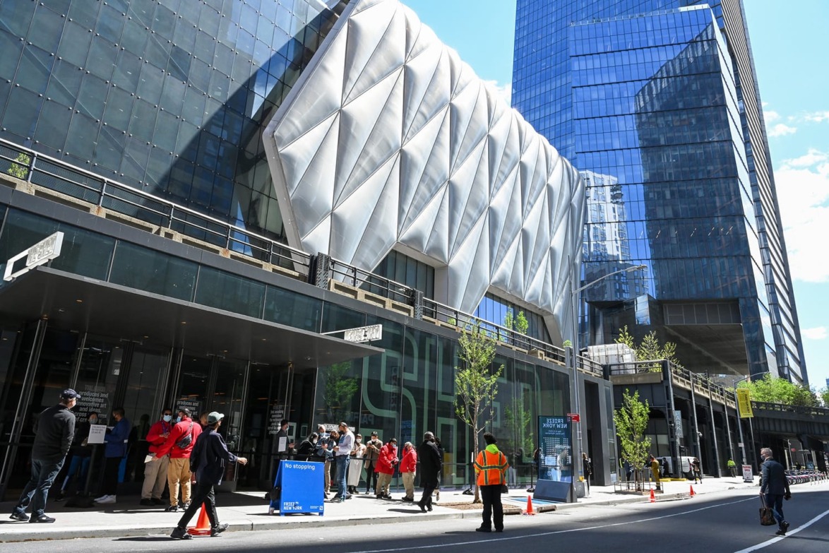 Modern building facade with geometric design, people lined up outside, and blue skies reflecting in glass windows.