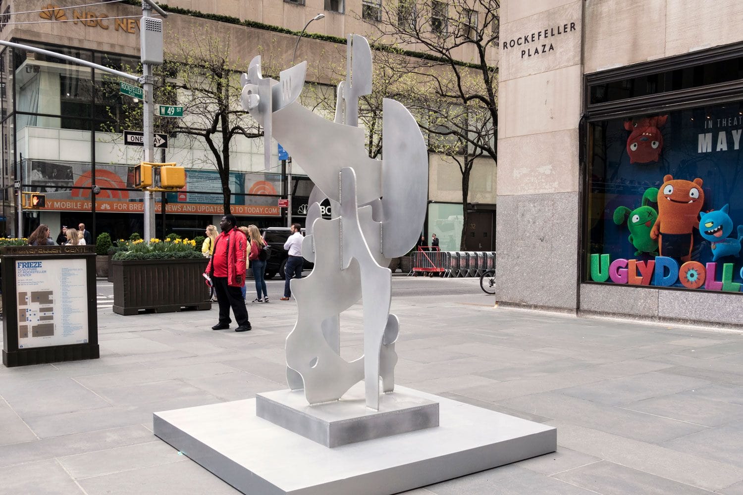 Artistic silver sculpture displayed outdoors in Rockefeller Plaza, surrounded by pedestrians and nearby storefronts.