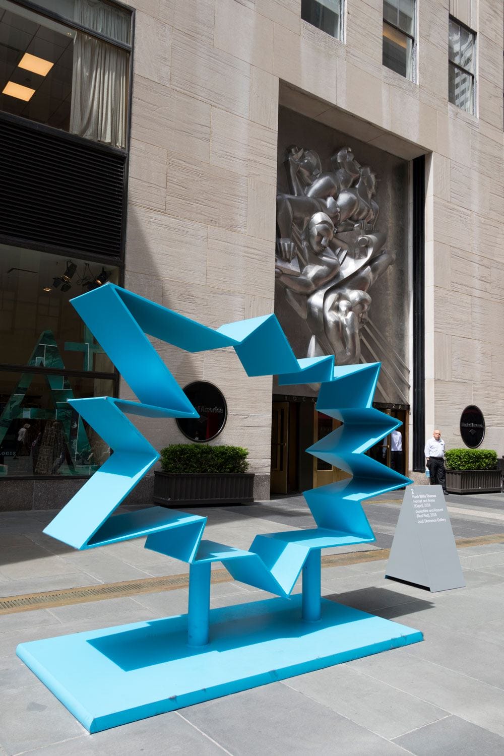 Blue abstract sculpture in front of a stone building with a decorative relief and glass doors.