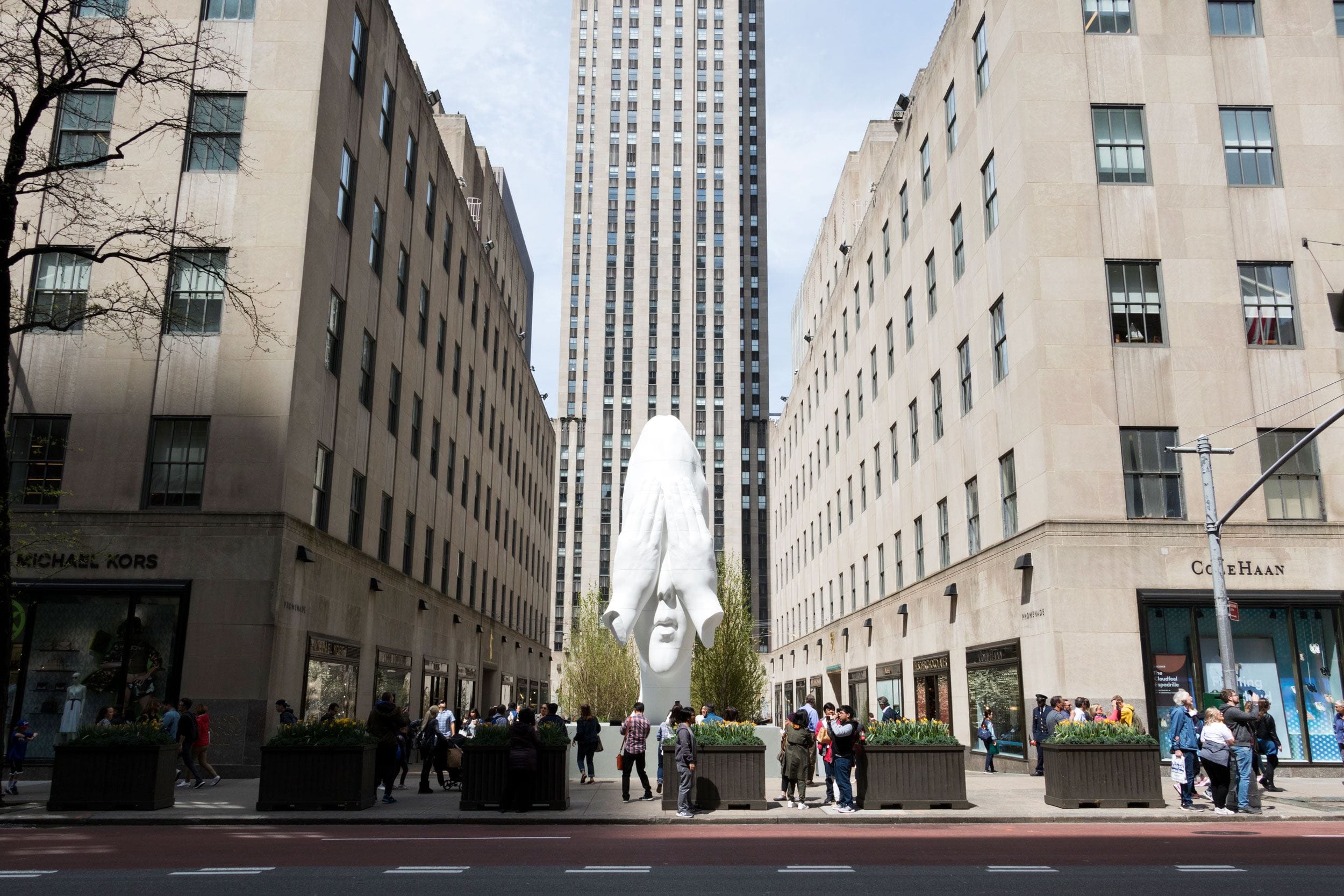 Large white head sculpture between buildings with people walking and a tall skyscraper in the background.