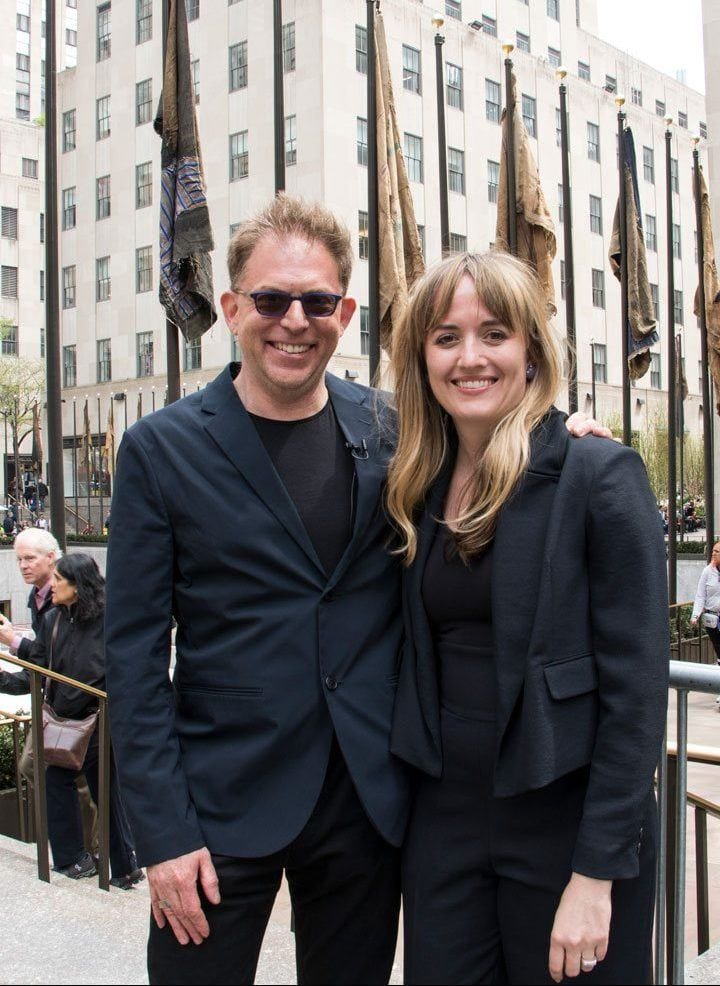 Man and woman smiling and standing together outdoors in an urban setting with flags and buildings in the background