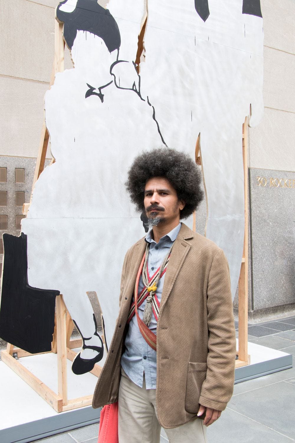 Man with an afro standing in front of a large abstract art piece outdoors.