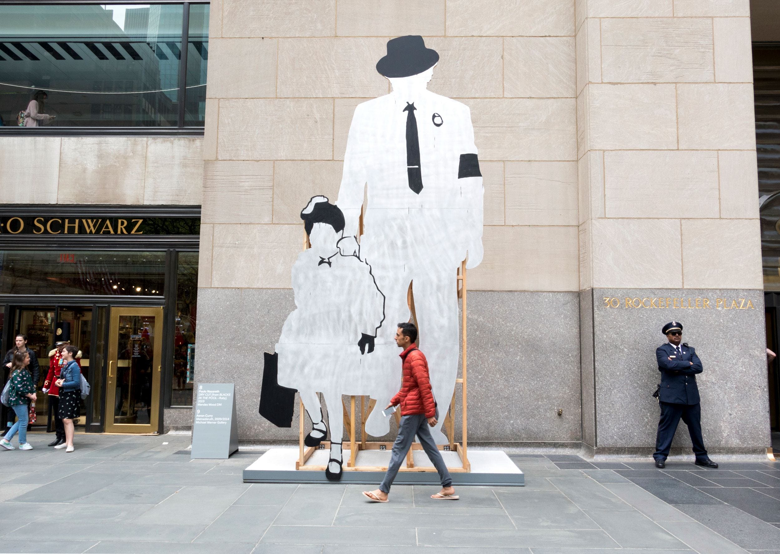 Sculpture of two faceless figures in business attire in front of FAO Schwarz store, with pedestrians passing by.