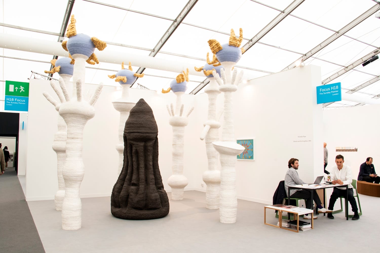 Art installation with tall white and black sculptures, blue accents, people seated and working at a table in an art fair setting.