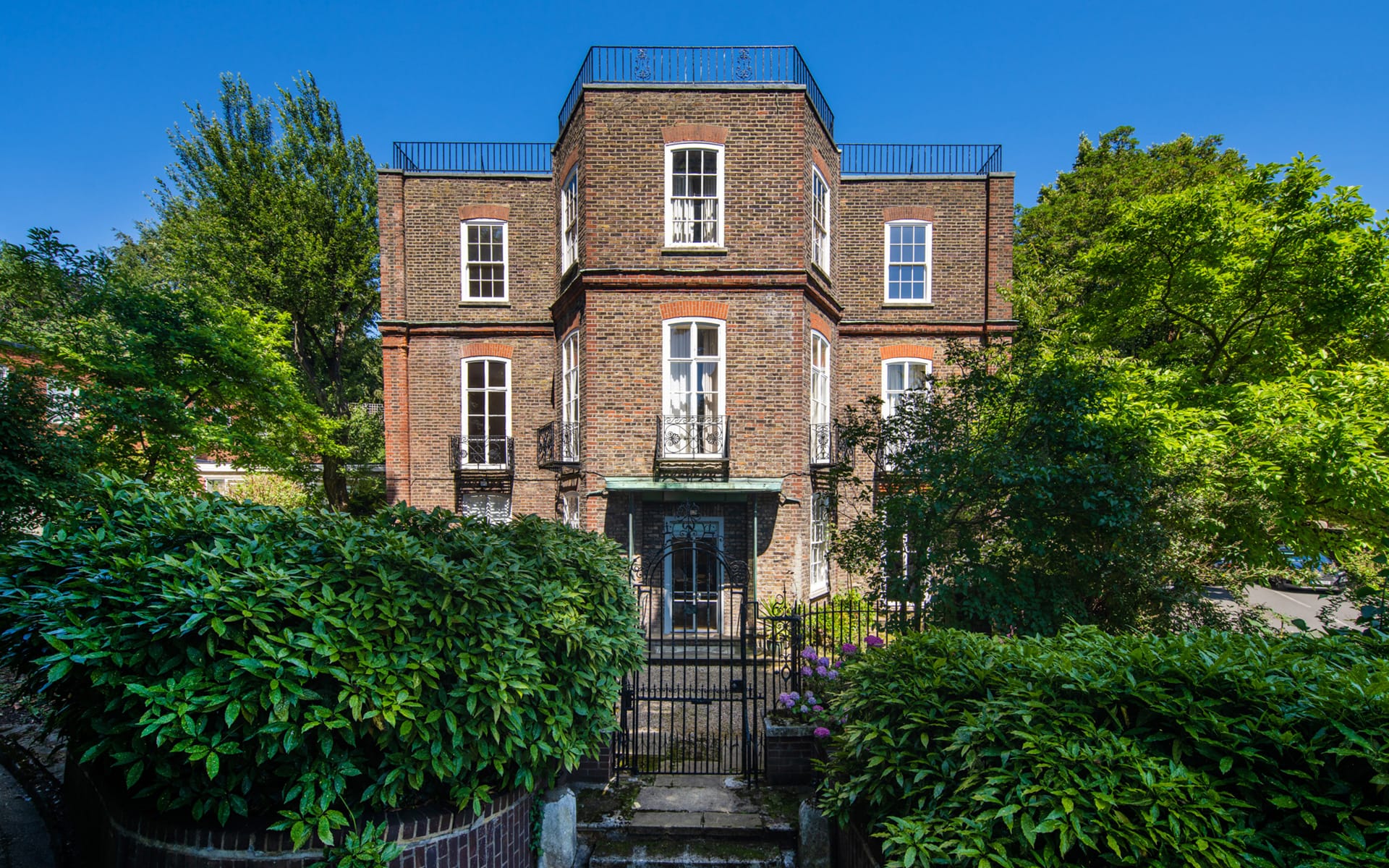 Three-story brick building surrounded by lush green trees and bushes on a bright sunny day.