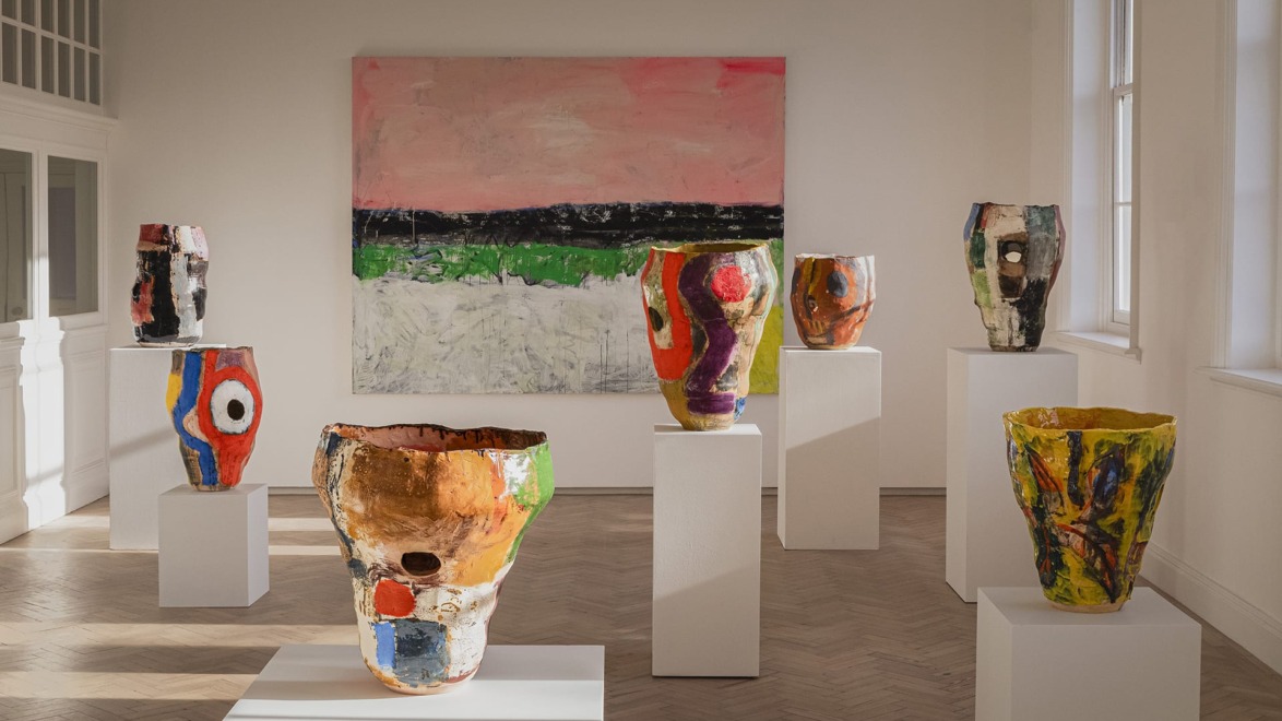 Colorful abstract ceramic vases on pedestals in an art gallery with a large painting on the wall in the background.