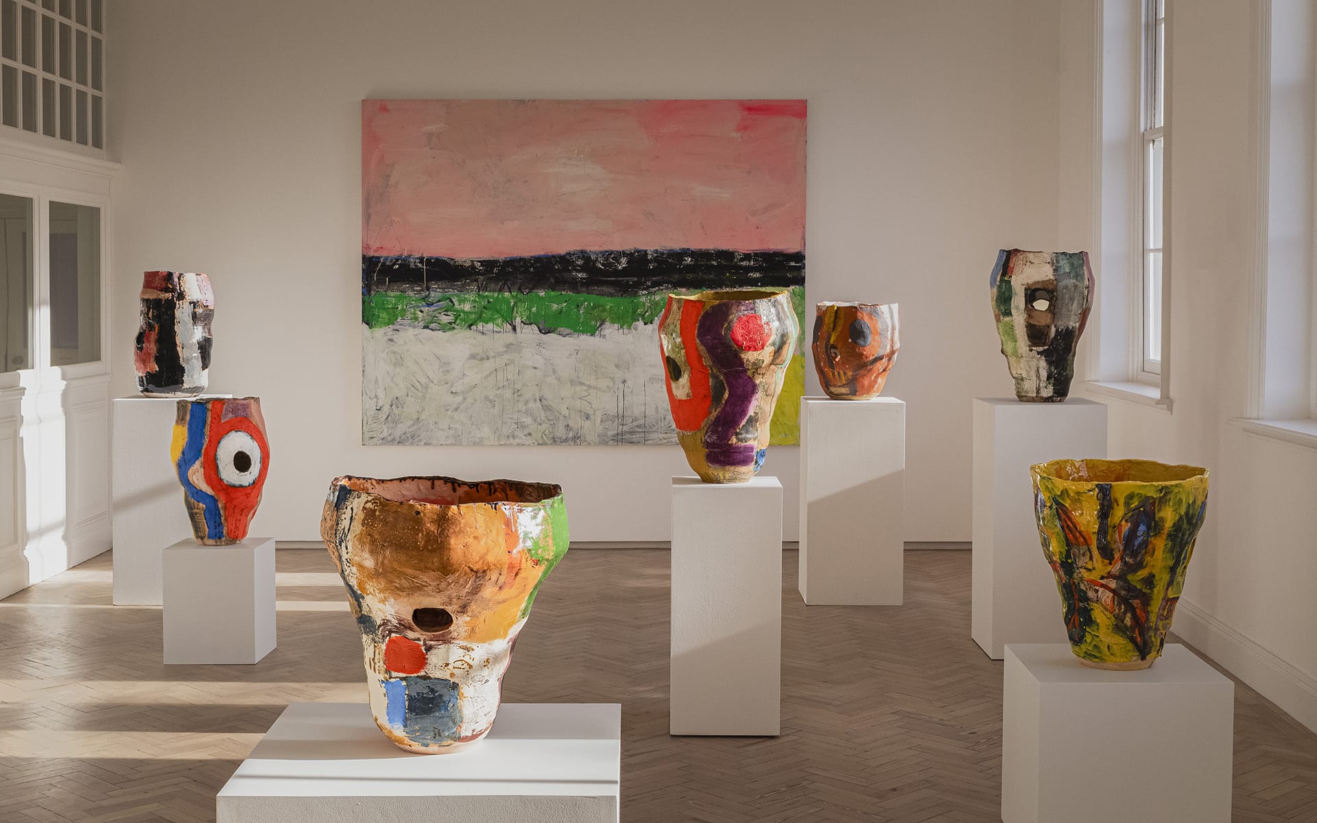 Colorful abstract ceramic vases on pedestals in an art gallery with a large painting on the wall in the background.