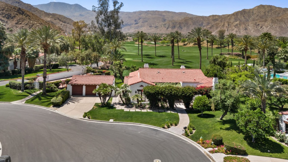 Aerial view of a villa with a red roof, surrounded by palm trees and mountains, near a golf course in a desert landscape.