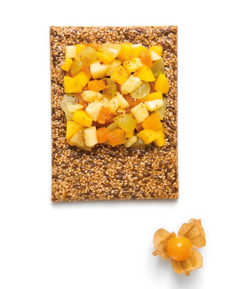 Colorful fruit-topped sesame brittle on white background with a small orange fruit beside it.