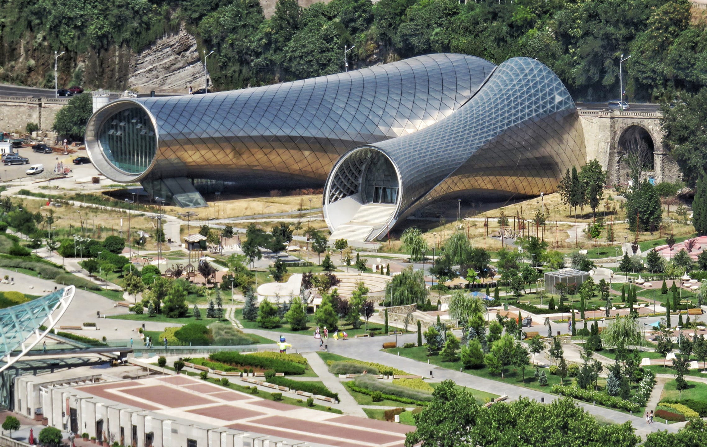 Modern architectural structure in a park with unique curved steel and glass design surrounded by greenery and pathways.