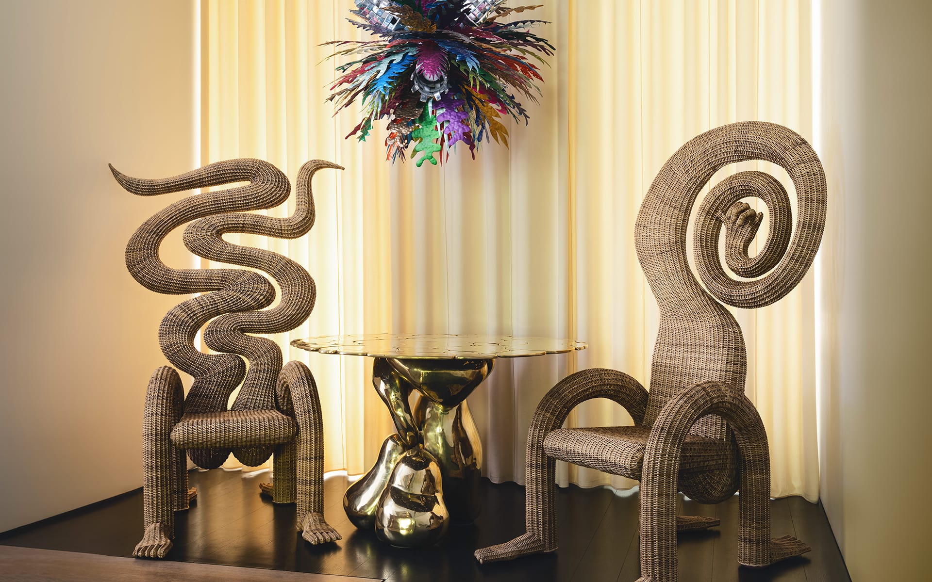 Two sculptural wicker chairs with abstract designs beside a round glass table with a gold base, under a colorful feather artwork.