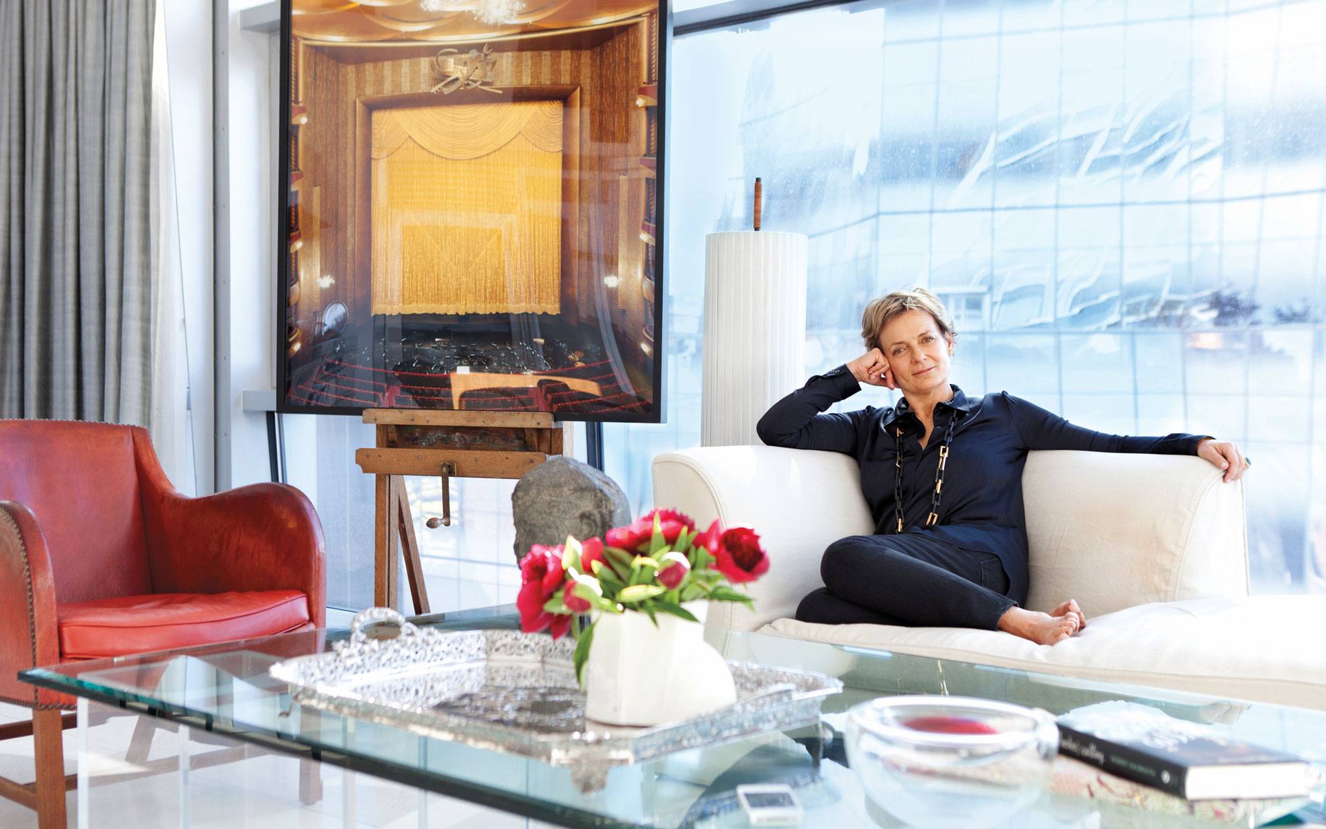Person sitting on a white sofa in a modern living room with a large painting and a vase of red flowers on the table.