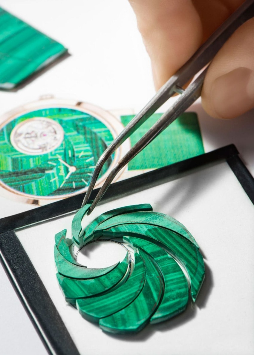 Close-up of green circular pattern being crafted with tweezers, with a watch face design in the background.