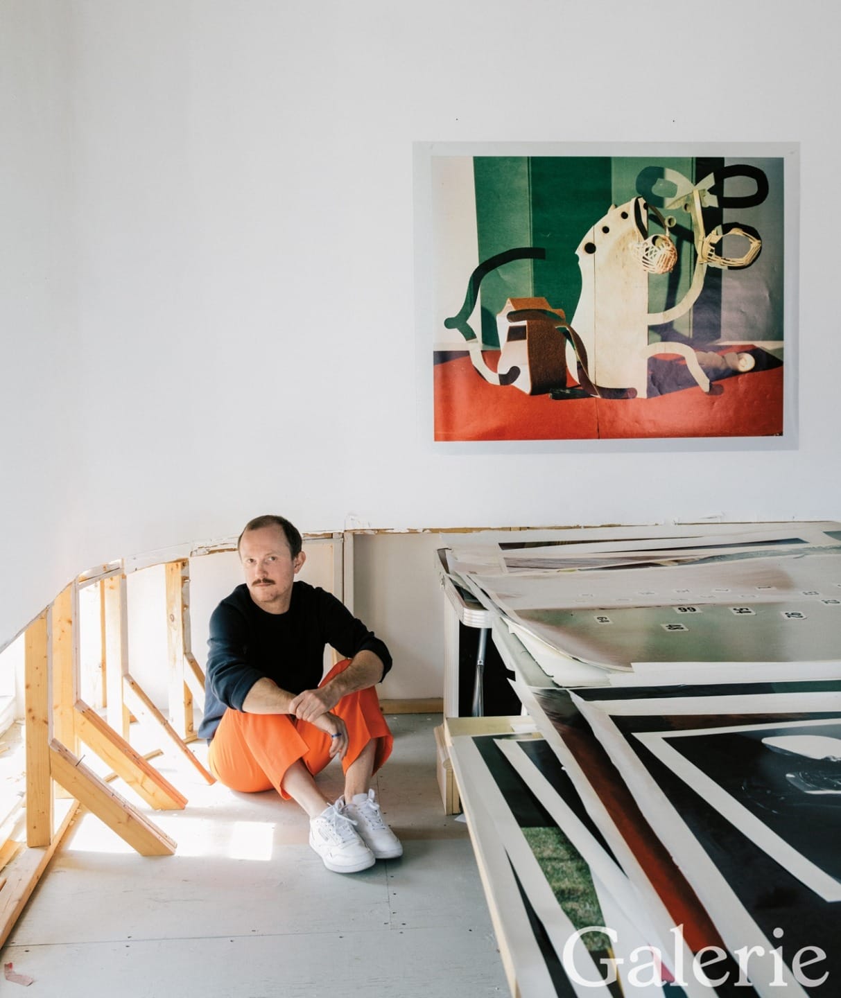 Artist sitting on floor in studio with colorful artwork on the wall and large prints on a table.