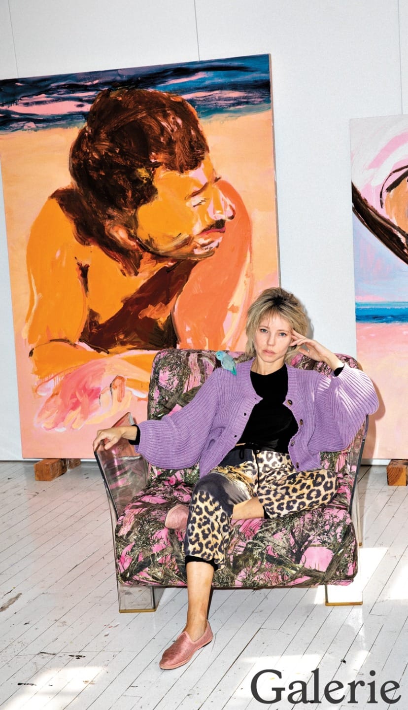 Person sitting in a chair wearing a purple sweater and leopard print pants, two colorful paintings in the background.