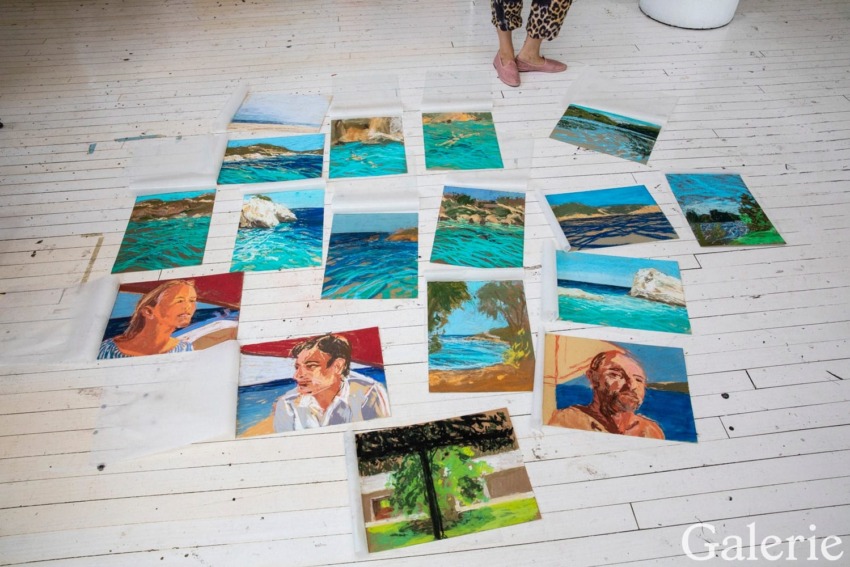 Several vibrant landscape and portrait paintings spread across a wooden floor with a person's legs visible at the top.