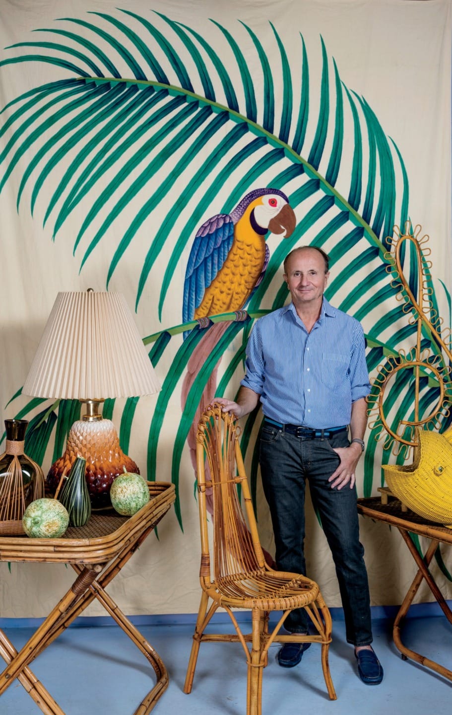 Man in blue shirt standing with rattan furniture, vibrant parrot mural, and tropical-themed decor in background.