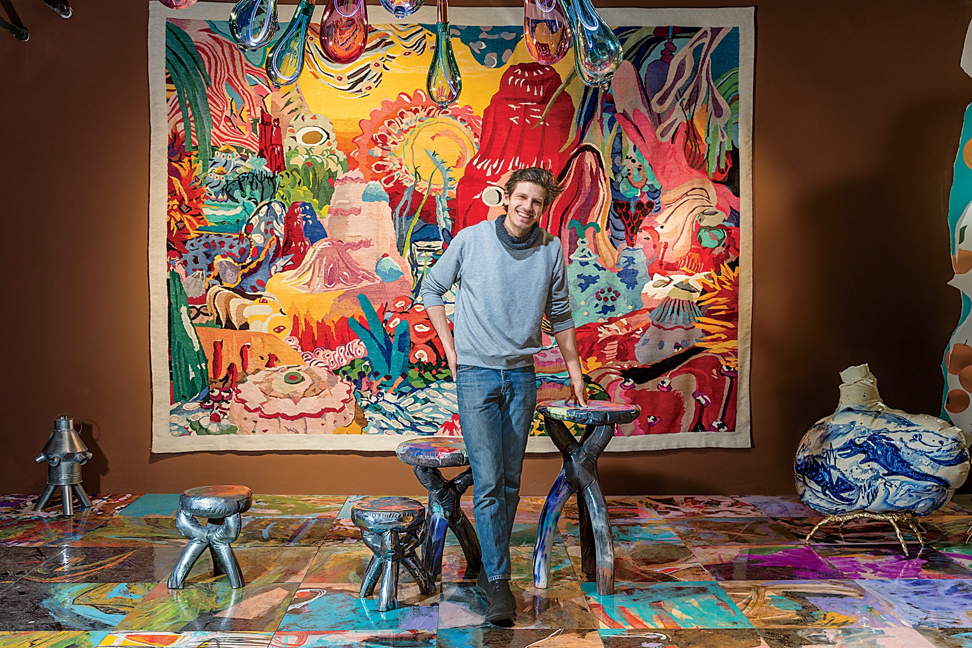 Person standing in front of a vibrant, abstract tapestry, with colorful floor art and uniquely shaped furniture.