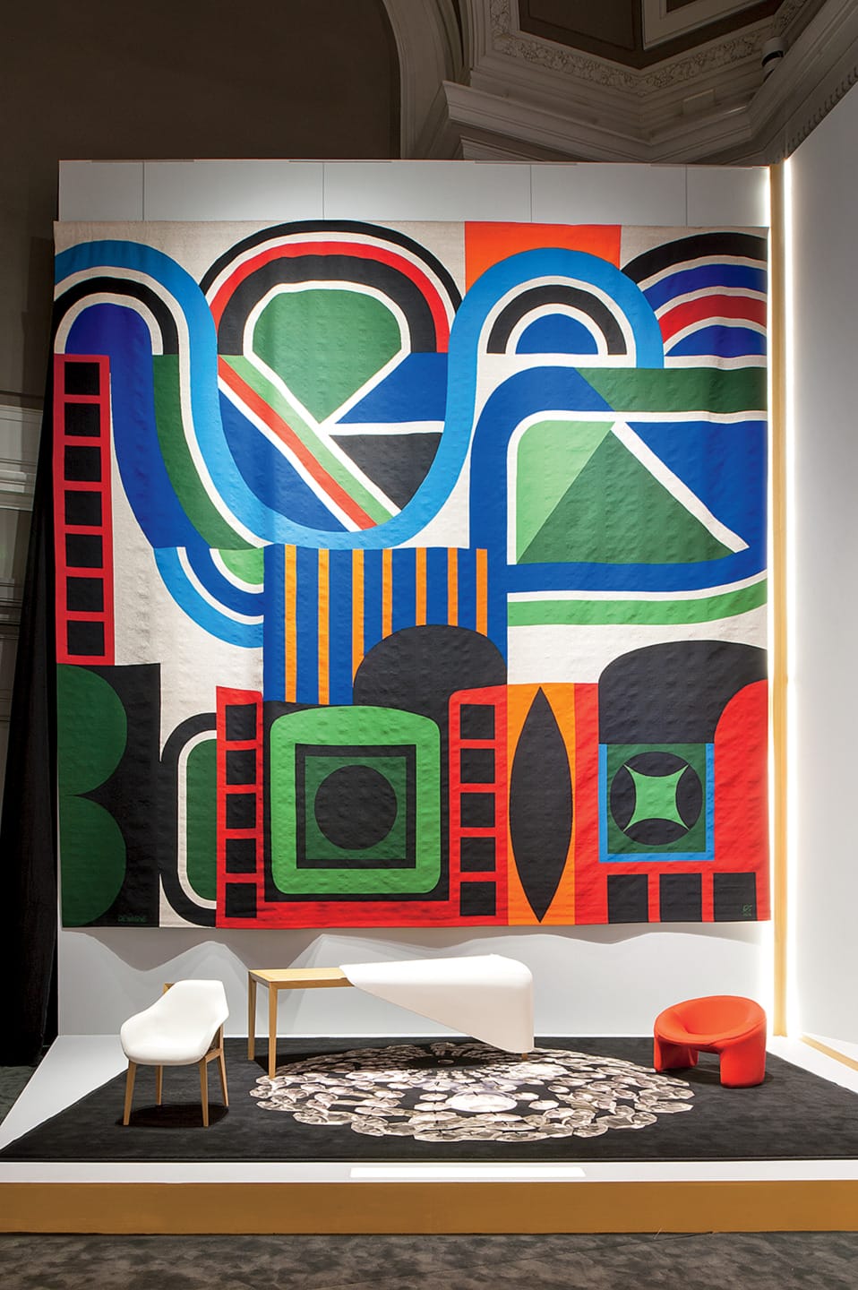 Colorful abstract art tapestry displayed on a wall above modern white and red chairs with a patterned rug.