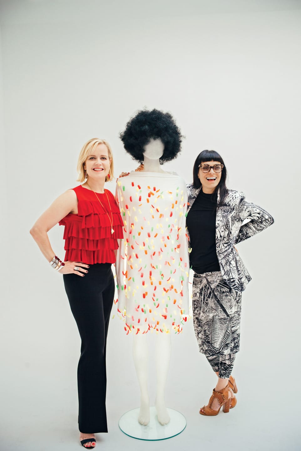 Two women posing with a mannequin dressed in a colorful outfit and a curly black wig against a plain white background.