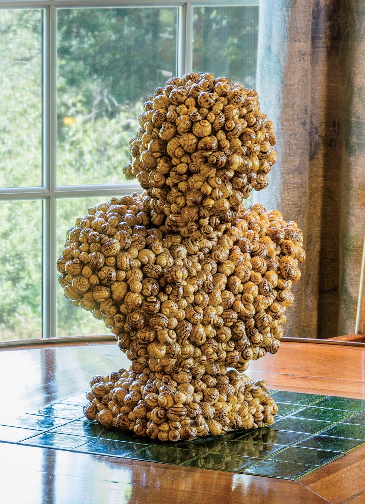 Bust sculpture made of numerous spiral shells on a wooden and tiled table near a window with nature view.