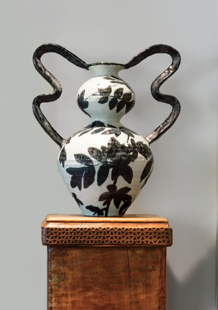 Ceramic vase with black floral design and ornate handles on a carved wooden pedestal against a gray background.