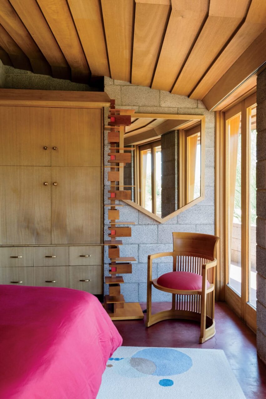 Bedroom with red bedspread, wooden chair, geometric mirror, wooden cabinetry, and large windows with natural light.