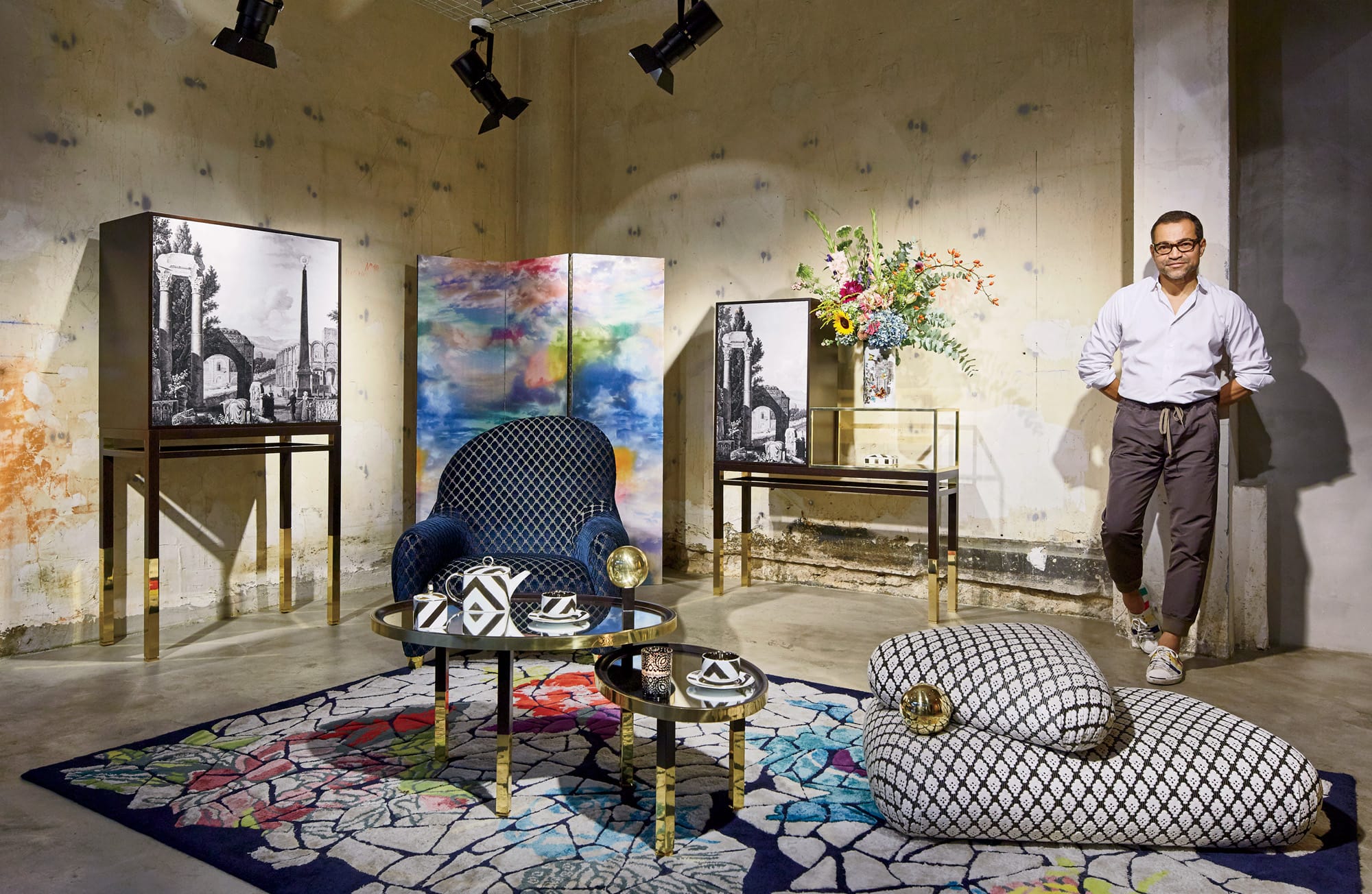 Designer standing in a stylish living room with modern furniture, colorful rug, artwork, and decorative pieces.
