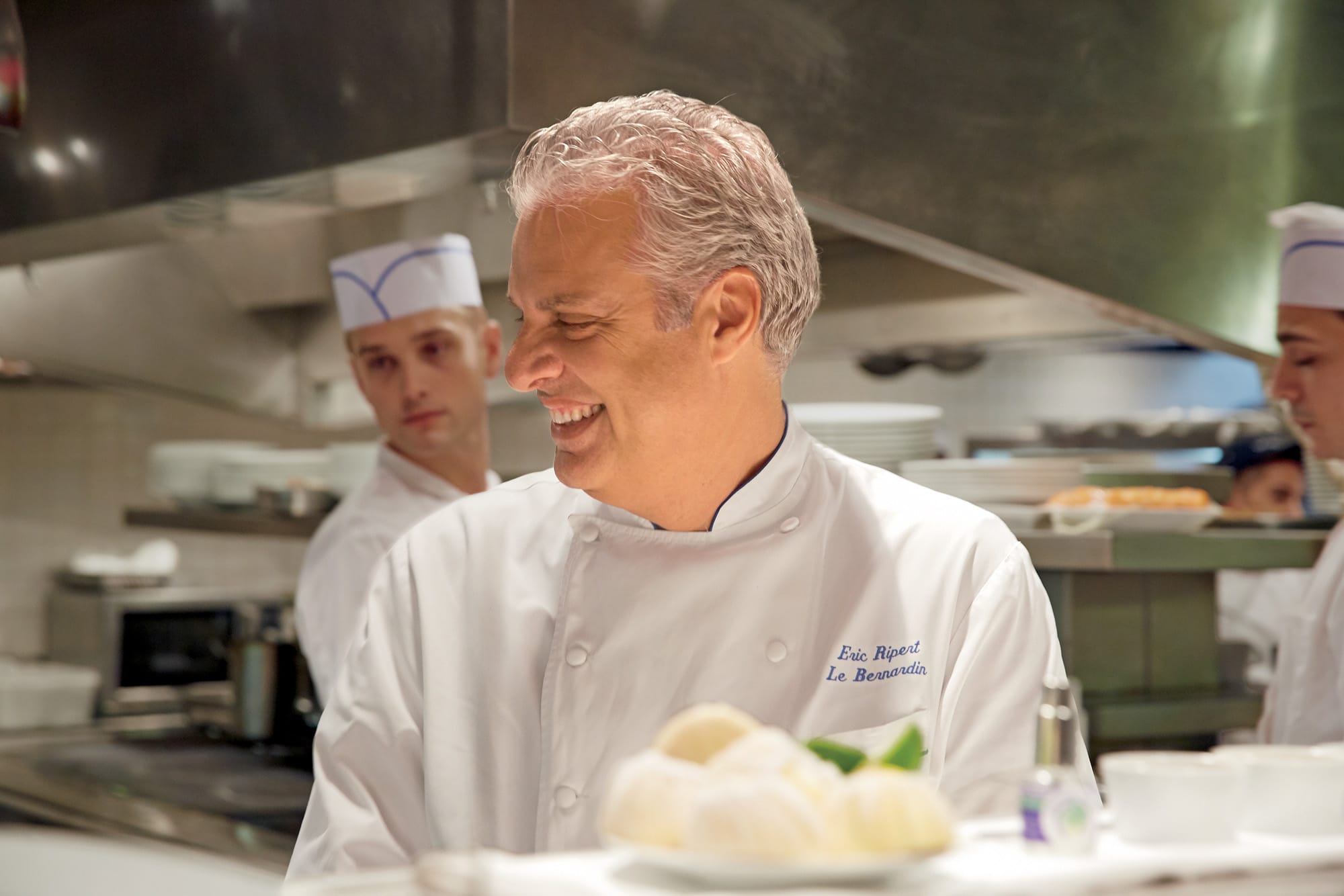 Chef smiling while standing in a busy kitchen, surrounded by staff members in white uniforms.