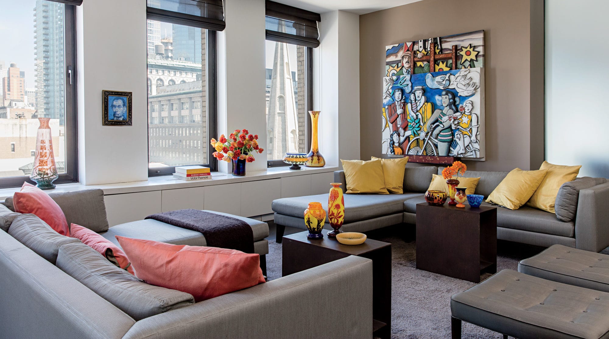Modern living room with large windows, gray sofas, colorful pillows, vibrant artwork, and decorative vases on tables.