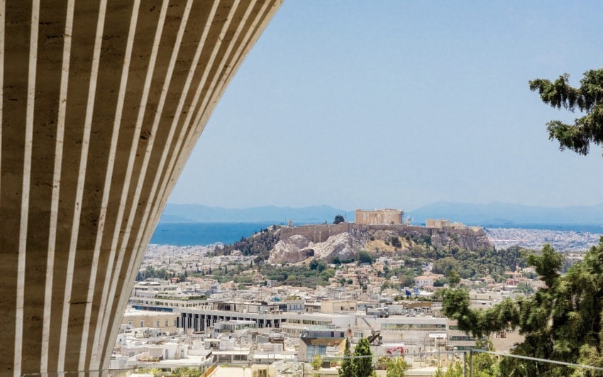Athens cityscape view with the Acropolis in the background, framed by modern architectural lines and trees in the foreground.