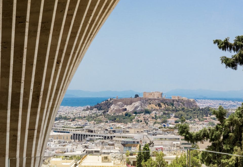 View of Athens cityscape framed by modern architectural structure, with historical ruins in the distance under clear blue sky