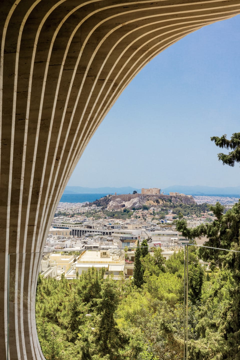 View of Athens cityscape framed by modern architectural structure, with historical ruins in the distance under clear blue sky